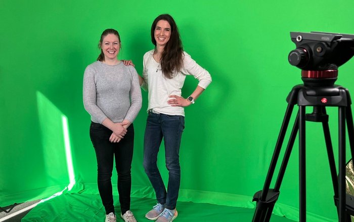 Zwei Frauen im Greenscreen Studio für das Streaming. Rechts vor den beiden steht ein Stativ für eine Kamera.