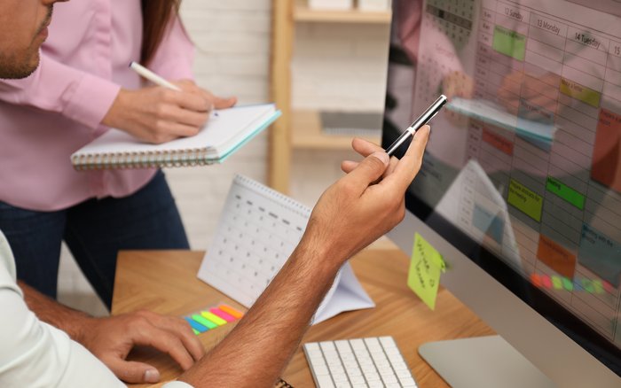 Zwei Personen sind an einem Schreibtisch. Eine Person zeigt mit dem Stift etwas im Kalender auf dem Rechner, die andere Person notiert etwas in ein Notizbuch.